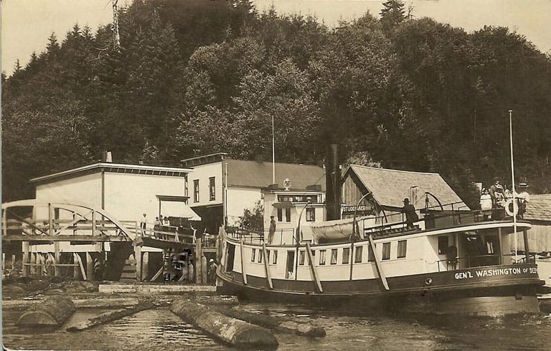 Black and white photo of the 'Gen'l. Washington' docked at Deep River. A group of ladies stand at the stern on the top deck. Two men are also on the top deck (one is leaning on the pilot house). The town of Deep River is visible in the background with four different buildings and storefronts including the Anderson Store along the bank of the river. One of the storefronts has a sign that reads 'Studebaker.'